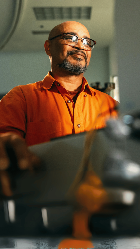 A person in glasses and an orange shirt operates a construction machine, smiling.