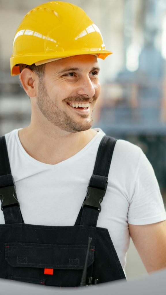 A person wearing a yellow hard hat and black overalls smiles while glancing to the side, embodying the spirit of construction.