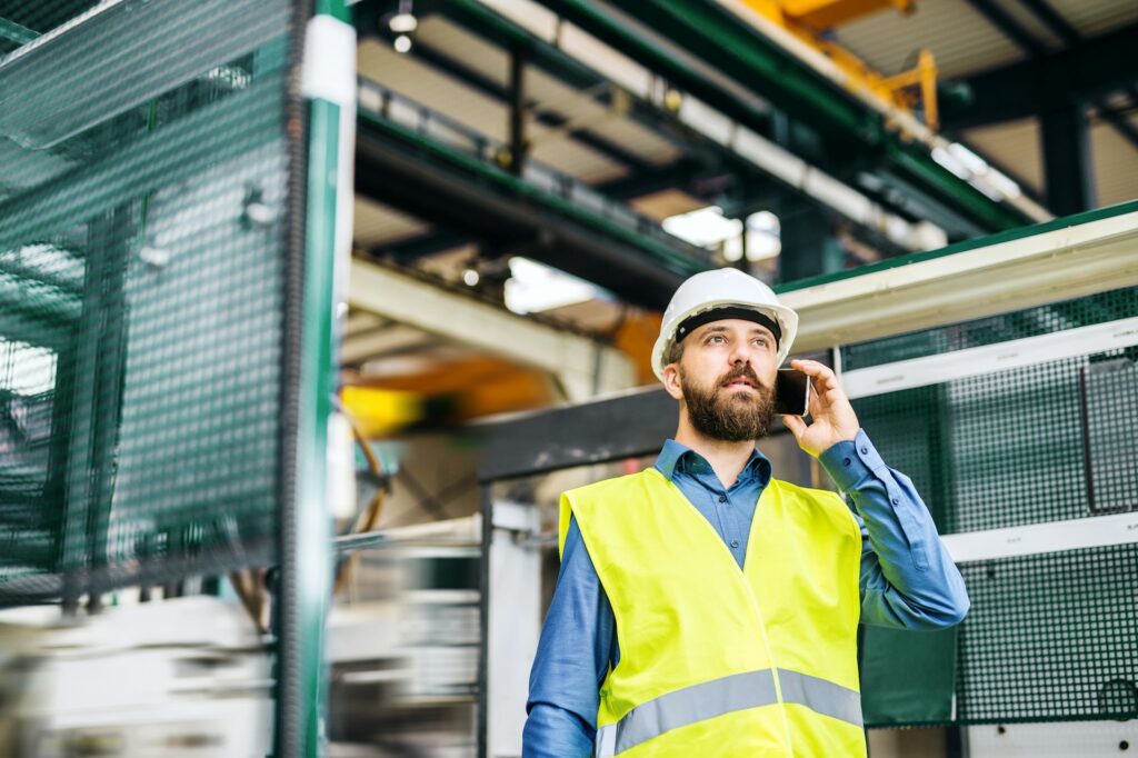 A man in a hard hat and safety vest talks on the phone in an industrial setting, discussing the latest updates in civil engineering recruitment.