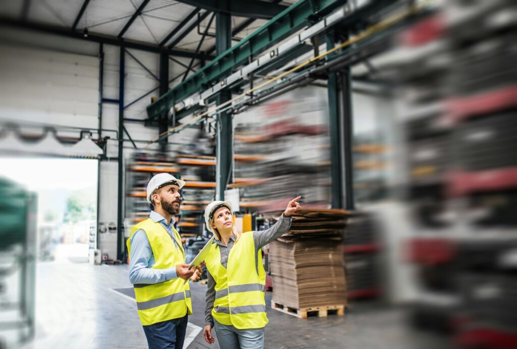 Two workers in safety vests and helmets stand in a warehouse, embodying the essence of construction. One points upward while the other holds a tablet, possibly reviewing geotechnical data. Pallets of goods are stacked in the background, creating a backdrop of organized efficiency.