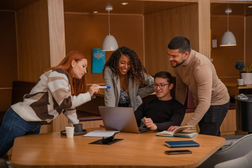 Four people are gathered around a table in an office, intently looking at a laptop screen. One person is gesturing towards the screen, with several books on geotechnical topics and a coffee cup scattered on the table, hinting at their focus on civil engineering recruitment strategies.