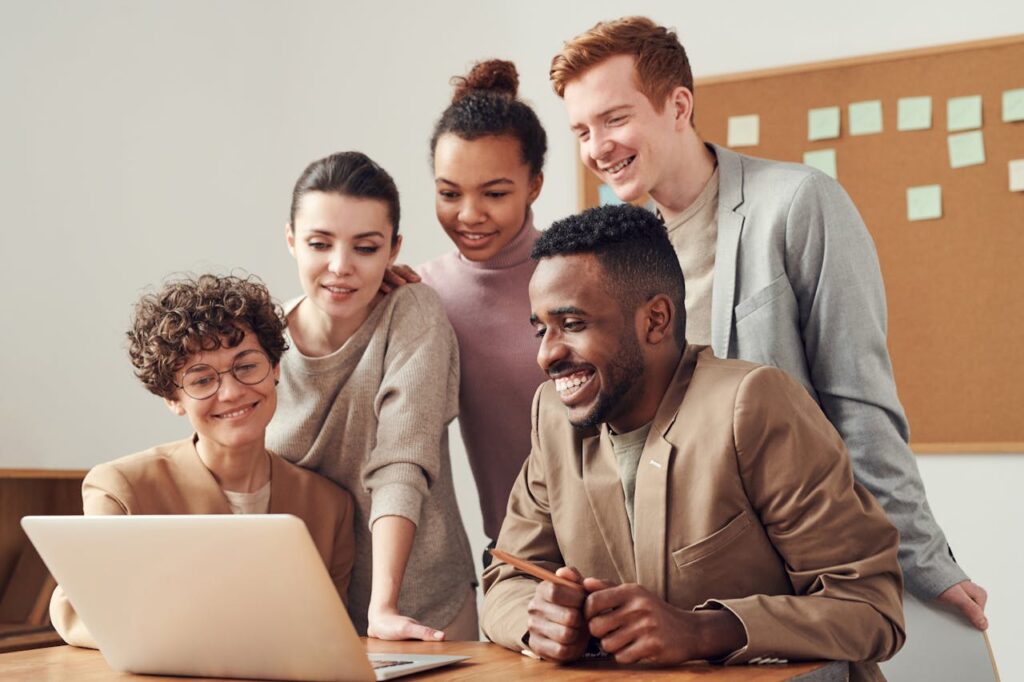 A group of five people gathers around a laptop, smiling and looking at the screen in an office setting. Behind them, a corkboard filled with construction project plans hints at a lively civil engineering recruitment discussion.